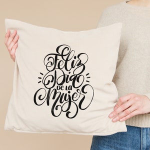 May include: A white cushion with black lettering that reads "Feliz Dia de la Mujer" in a cursive font.