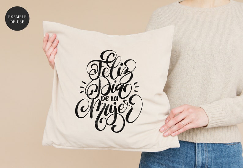 May include: A white cushion with black lettering that reads "Feliz Dia de la Mujer" in a cursive font.