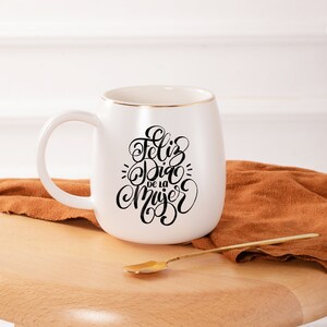 May include: White ceramic mug with a gold rim and black calligraphy that reads "Feliz Dia de la Mujer". The mug is sitting on a wooden surface with a brown fabric next to it.