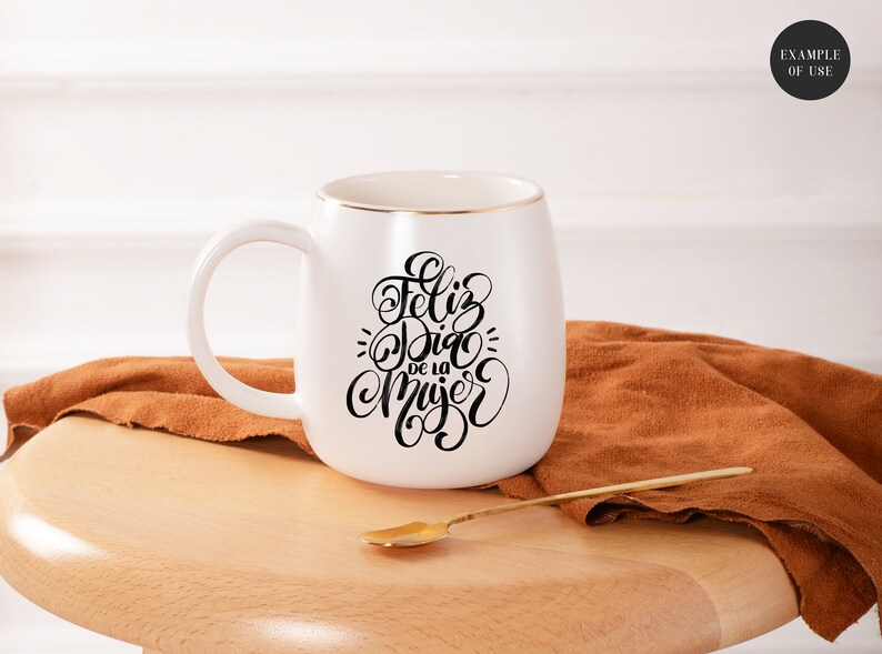 May include: White ceramic mug with a gold rim and black calligraphy that reads "Feliz Dia de la Mujer". The mug is sitting on a wooden surface with a brown fabric next to it.