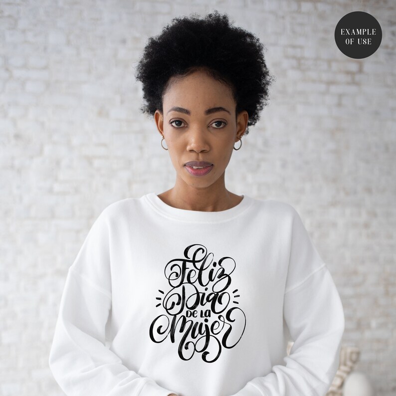May include: White sweatshirt with black lettering that says "Feliz Dia de la Mujer" which translates to "Happy Women's Day" in English.