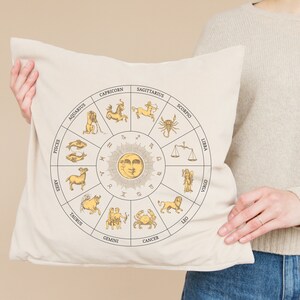 Op de afbeelding: Een wit kussen met een beige ontwerp van een zodiac-wiel met de 12 astrologische tekens en een zon met een gezicht. Het kussen wordt vastgehouden door een persoon die een cr&egrave;mekleurige trui en een blauwe jeans draagt.