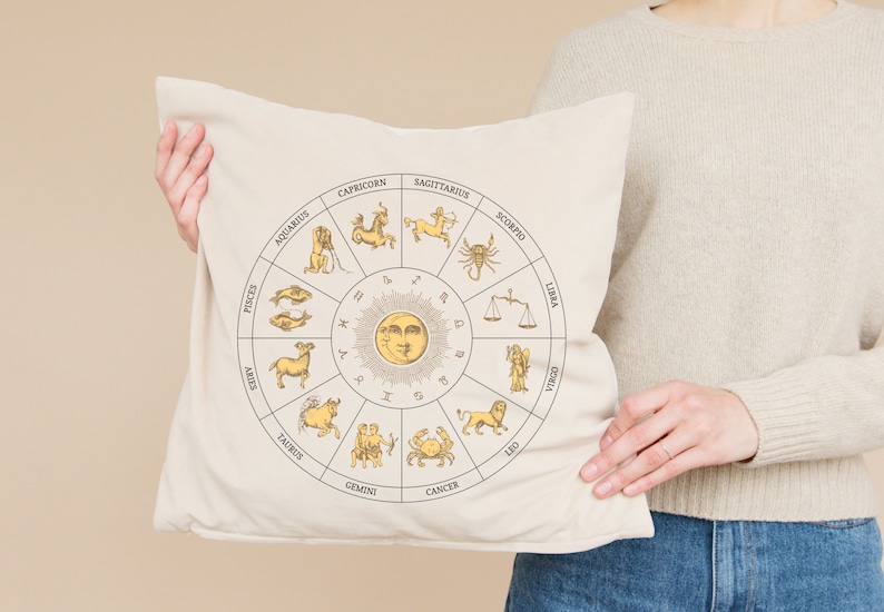Op de afbeelding: Een wit kussen met een beige ontwerp van een zodiac-wiel met de 12 astrologische tekens en een zon met een gezicht. Het kussen wordt vastgehouden door een persoon die een cr&egrave;mekleurige trui en een blauwe jeans draagt.