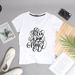 May include: White v-neck t-shirt with black lettering that says "Feliz Dia de la Mujer" in a cursive font.