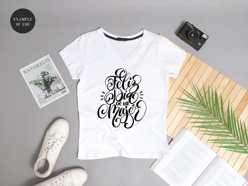 May include: White v-neck t-shirt with black lettering that says "Feliz Dia de la Mujer" in a cursive font.