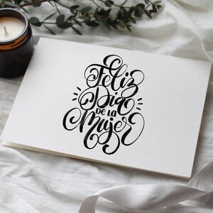 May include: A white card with black calligraphy that reads "Feliz Dia de la Mujer" which translates to "Happy Women's Day" in English.