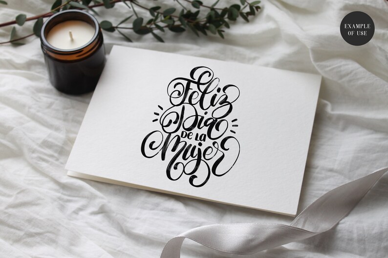 May include: A white card with black calligraphy that reads "Feliz Dia de la Mujer" which translates to "Happy Women's Day" in English.