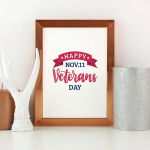 Puede incluir: Una impresi&oacute;n enmarcada con un gr&aacute;fico rojo y azul que dice "Happy Nov. 11 Veterans Day" con una cinta roja y estrellas. La impresi&oacute;n est&aacute; en un marco de cobre y est&aacute; sentada sobre una superficie blanca con una asta de ciervo blanca y dos velas.