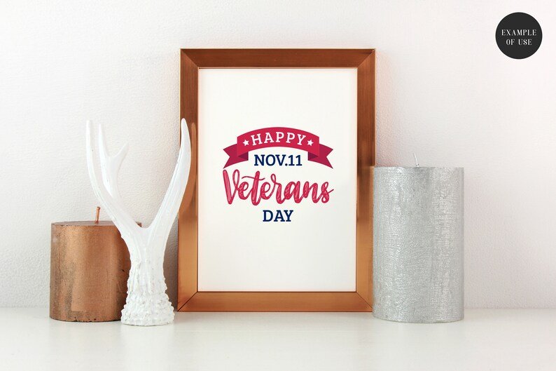Puede incluir: Una impresi&oacute;n enmarcada con un gr&aacute;fico rojo y azul que dice "Happy Nov. 11 Veterans Day" con una cinta roja y estrellas. La impresi&oacute;n est&aacute; en un marco de cobre y est&aacute; sentada sobre una superficie blanca con una asta de ciervo blanca y dos velas.