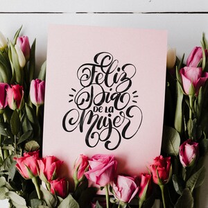 May include: A pink card with black calligraphy that reads "Feliz Dia de la Mujer" surrounded by pink and white tulips and roses.