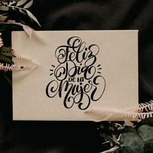 May include: A brown card with the text "Feliz Dia de la Mujer" written in black calligraphy.