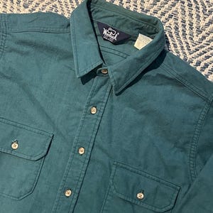 May include: Teal-colored button-down shirt with two chest pockets and a collar. The shirt has a button-up front and buttons on the cuffs. The Woolrich logo is visible on the collar. The shirt is made of a soft, possibly flannel, material.