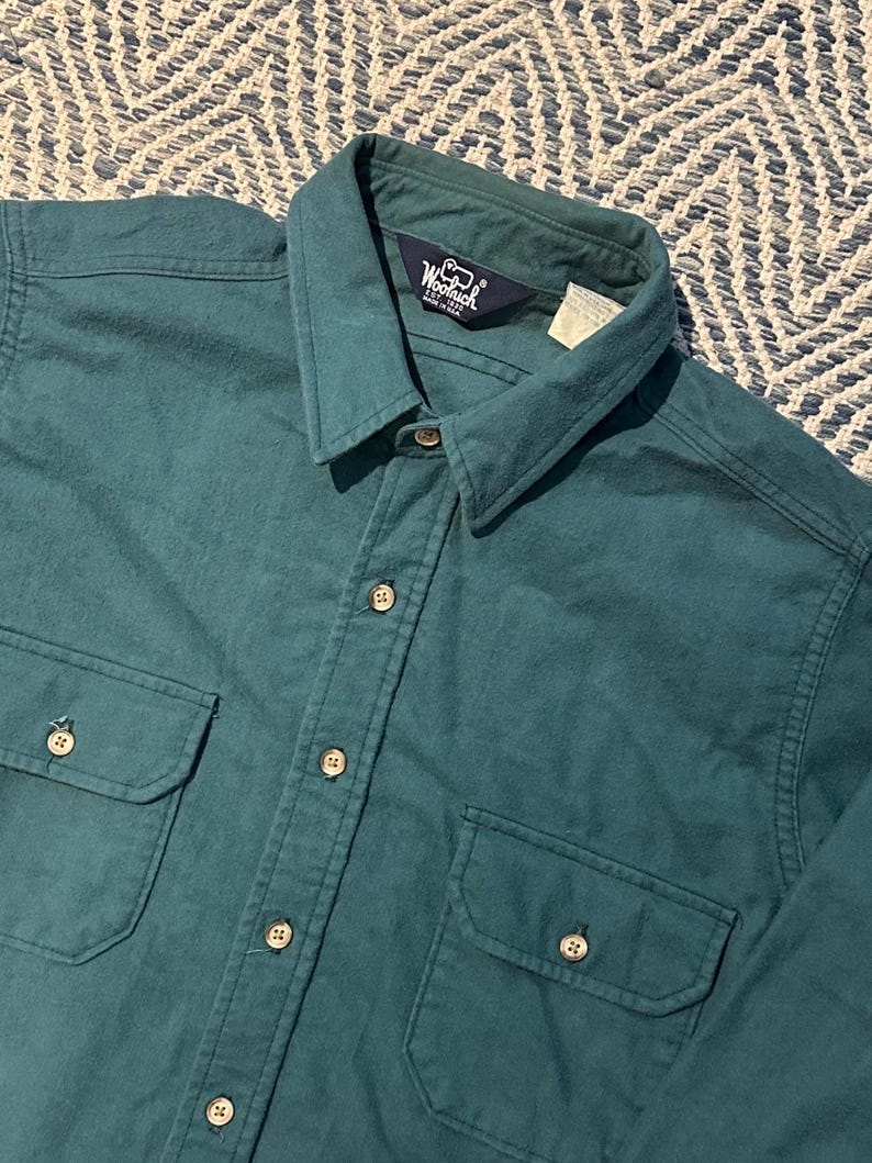 May include: Teal-colored button-down shirt with two chest pockets and a collar. The shirt has a button-up front and buttons on the cuffs. The Woolrich logo is visible on the collar. The shirt is made of a soft, possibly flannel, material.