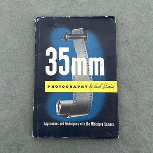 Puede incluir: Una cubierta de libro azul y amarilla con el título "35mm FOTOGRAFÍA por Jacob Deschin" y el subtítulo "Enfoques y técnicas con la cámara en miniatura". La cubierta presenta un gráfico de un rollo de película.