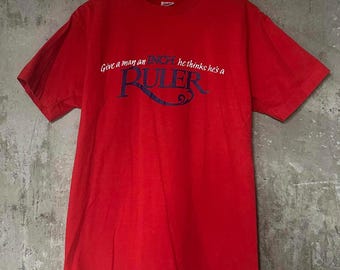 May include: A red t-shirt with the text "Give a man an inch, he thinks he's a RULER" in white and blue. The shirt is hanging on a wooden hanger against a textured gray background. The t-shirt is a crew neck style.