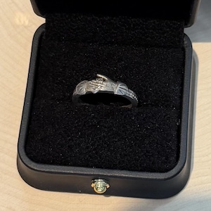 May include: A silver-toned ring featuring a design of two hands clasping each other, presented inside a black square jewelry box with a black velvet interior lining. The ring band displays detailed engravings on both sides of the central hand motif.