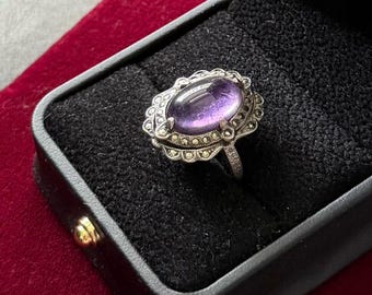 Puede incluir: Un anillo de plata con una piedra amatista morada en forma de cabujón ovalado en el centro. El engaste está rodeado por un borde decorativo de pequeñas piedras de marcasita redondas y gris oscuro dispuestas en un patrón metálico festoneado.