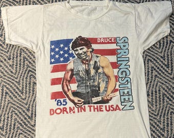 May include: White t-shirt featuring a graphic of Bruce Springsteen playing a guitar, with an American flag design and the text "BRUCE SPRINGSTEEN '85 BORN IN THE USA". The shirt has short sleeves and a crew neck.