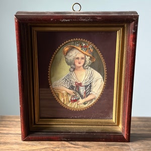 May include: A framed portrait of a woman wearing a straw hat with flowers. The portrait is set against a burgundy background and has a gold frame.