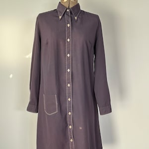 May include: A long-sleeved, dark purple button-down shirt dress with a single pocket on the left side. The dress has white stitching around the edges of the collar, cuffs, and pocket.