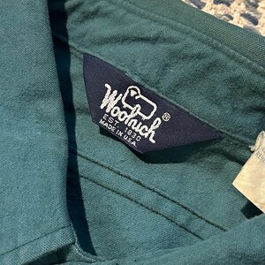 May include: Close-up of a teal-colored shirt collar with a navy blue label. The label reads "Woolrich EST. 1830 MADE IN U.S.A." in white lettering. The shirt is made of a textured fabric and has a button closure.