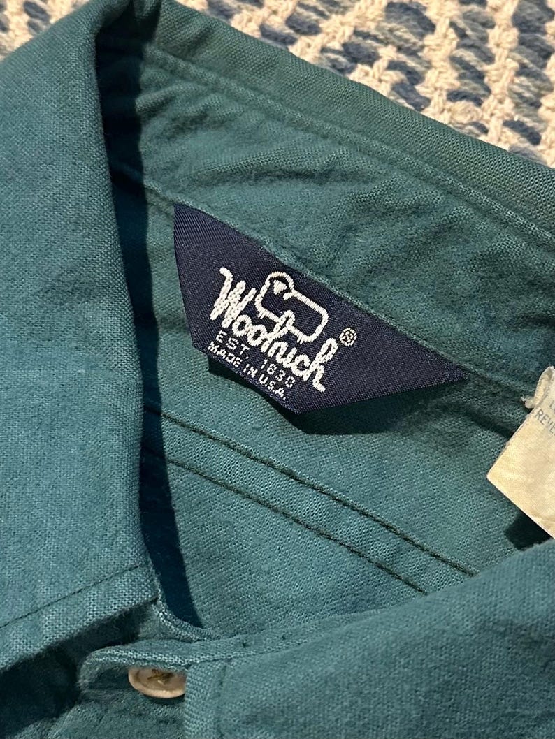 May include: Close-up of a teal-colored shirt collar with a navy blue label. The label reads "Woolrich EST. 1830 MADE IN U.S.A." in white lettering. The shirt is made of a textured fabric and has a button closure.