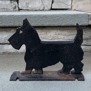 May include: A black metal Scottie dog doorstop. The doorstop is a silhouette of a Scottish Terrier, with a rectangular base and a slightly rusted appearance. The dog has a pointed ear and a raised tail.