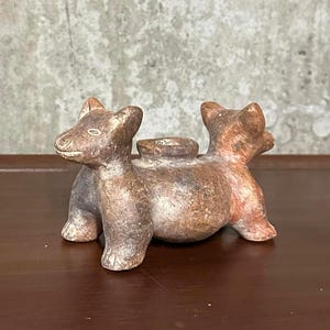 Pode incluir: Uma escultura de cerâmica marrom de um animal de duas cabeças com uma pequena abertura circular nas costas. A escultura tem uma superfície áspera e texturizada e está sobre um fundo neutro.