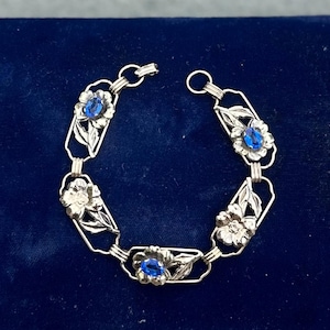 May include: Silver-toned bracelet featuring floral designs with blue gemstone centers. The bracelet has an open link design with detailed leaves and flowers, set against a dark blue background.