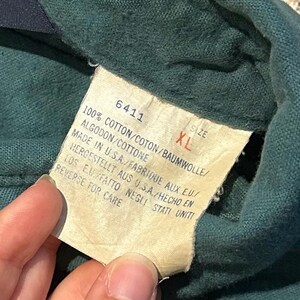 May include: Close-up of a green garment with a tag. The tag displays the number 6411, size XL, and details about the fabric composition: 100% cotton. It also indicates the garment was made in the USA.