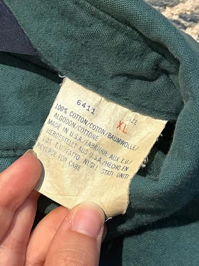 May include: Close-up of a green garment with a tag. The tag displays the number 6411, size XL, and details about the fabric composition: 100% cotton. It also indicates the garment was made in the USA.