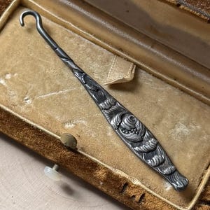 May include: An antique silver crochet hook with ornate floral detailing, resting inside a vintage velvet-lined case. The hook features a curved end and a decorative handle. The case is a warm brown color.