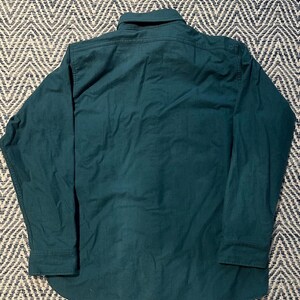 May include: A dark teal long-sleeve shirt with a button-down collar and cuffs. The shirt is made of a solid-colored fabric and has a curved hemline. The back of the shirt is visible, showing the collar and the yoke.