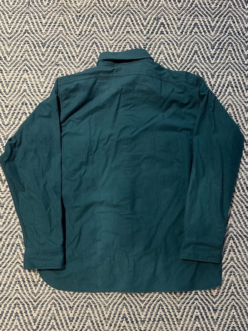 May include: A dark teal long-sleeve shirt with a button-down collar and cuffs. The shirt is made of a solid-colored fabric and has a curved hemline. The back of the shirt is visible, showing the collar and the yoke.