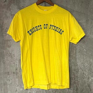 Könnte beinhalten: Ein leuchtend gelbes T-Shirt mit dem Text "KNIGHTS OF PYTHIAS" in Marineblau, der bogenförmig über der Brust angeordnet ist. Das T-Shirt hängt an einem Holzbügel vor einem strukturierten grauen Hintergrund. Es hat kurze Ärmel und einen Rundhalsausschnitt.