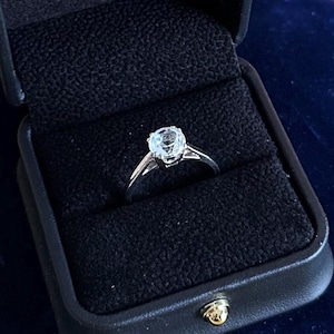 May include: A silver engagement ring with a large, clear gemstone is displayed inside a black ring box. The ring is centered in the box, which is lined with black fabric. The box is closed and set against a dark blue background.