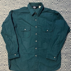 May include: A long-sleeved, teal-colored button-down shirt with two chest pockets. The shirt has a classic collar and buttoned cuffs. The fabric appears to be a durable, medium-weight material.