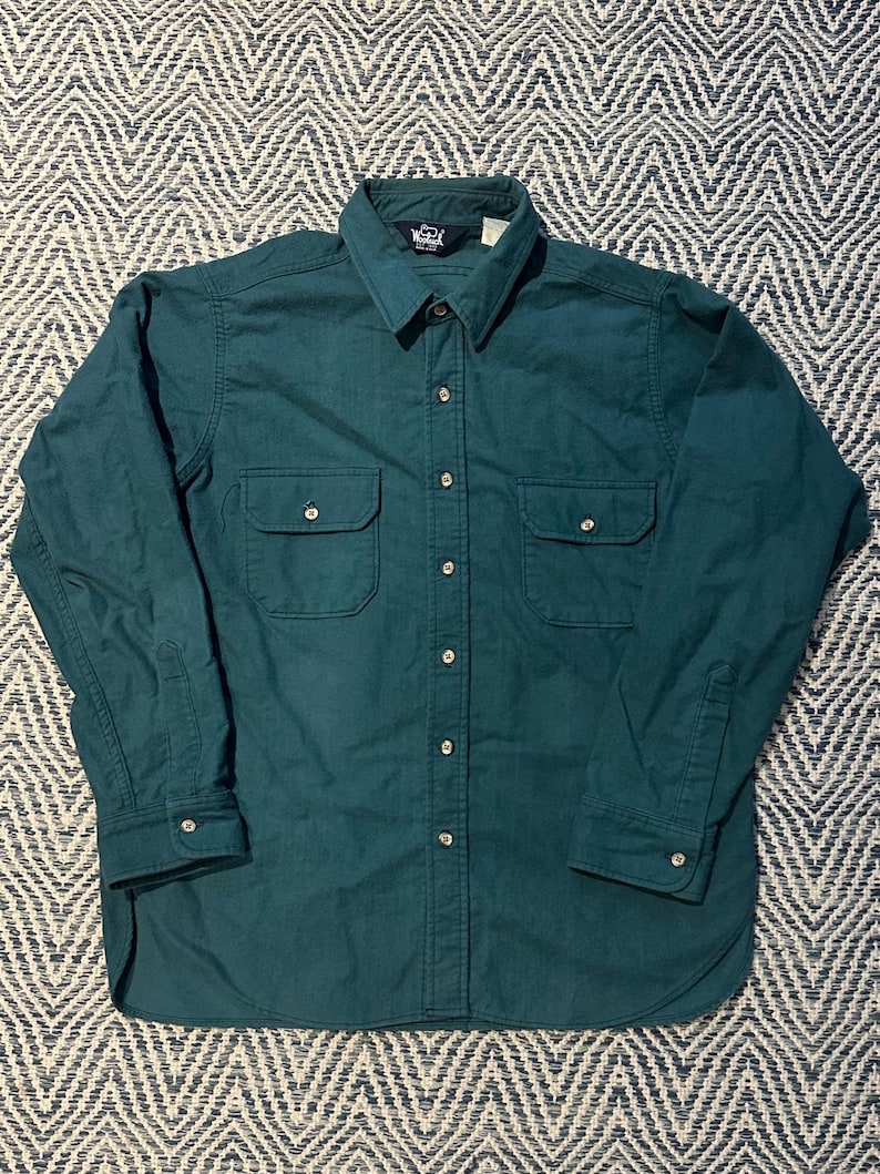 May include: A long-sleeved, teal-colored button-down shirt with two chest pockets. The shirt has a classic collar and buttoned cuffs. The fabric appears to be a durable, medium-weight material.