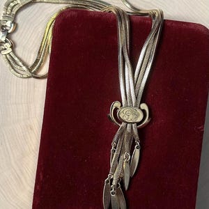 May include: A gold-toned necklace with multiple strands and a decorative pendant. The pendant features an oval shape with an engraved design and dangling leaf-shaped charms. The necklace is displayed on a red velvet surface.