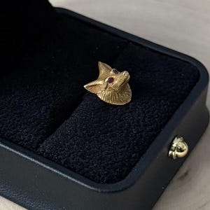 May include: A gold-colored fox head pin with red gemstone eyes, displayed in a black jewelry box. The pin is detailed with textured fur and a pointed snout. The box has a black velvet interior and a gold clasp.