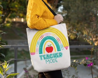 May include: A white tote bag with a rainbow design made of pencils, paper clips, and a ruler. The rainbow has a red apple in the center. The text "Teacher Mode" is printed below the rainbow.