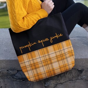 Peut inclure: Un sac fourre-tout noir avec un motif à carreaux jaune et blanc sur la moitié inférieure. Le sac porte le texte "pumpkin spice junkie" écrit en orange sur la partie noire.