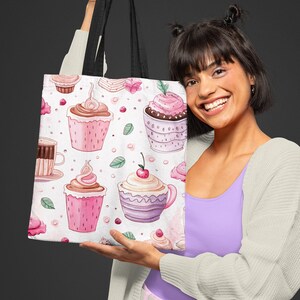 Op de afbeelding: Een witte tote bag met een zwarte handvat heeft een herhalend patroon van roze en paarse cupcakes met kersen en koffiekopjes. De tas wordt vastgehouden door een persoon die een lichtgrijze cardigan, een lavendelkleurig tanktop en een roze broek draagt.