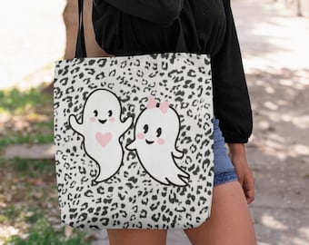 May include: A white tote bag with a black and white leopard print background. Two cartoon ghosts are featured on the front, one with a pink heart and the other with a pink bow. The bag is carried over the shoulder.