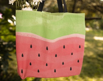 May include: A green and red tote bag with a watermelon design. The bag has black straps and is hanging from a tree branch. A bouquet of white daisies is inside the bag.