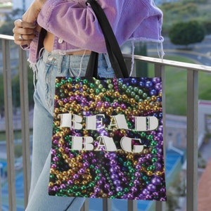 May include: A tote bag with a colorful bead design in shades of purple, gold, and green. The words "BEAD BAG" are printed in white block letters on the front. The bag has black straps and is held by a person wearing a purple denim jacket.