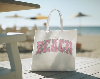 May include: White canvas tote bag with pink lettering that says "BEACH". The bag is sitting on a wooden surface.