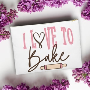May include: White canvas zippered pouch with the text "I love to bake" in pink lettering with a brown heart and a brown rolling pin graphic.