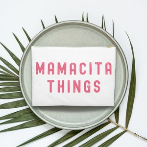 May include: A white zippered pouch with the text "MAMACITA THINGS" printed in pink on a gray ceramic plate with green palm leaves on a white background.