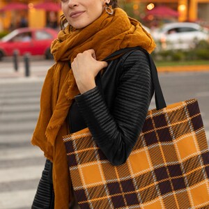 May include: A woman is carrying a large tote bag with a black and orange plaid pattern. The bag has black handles and is made of a canvas material.